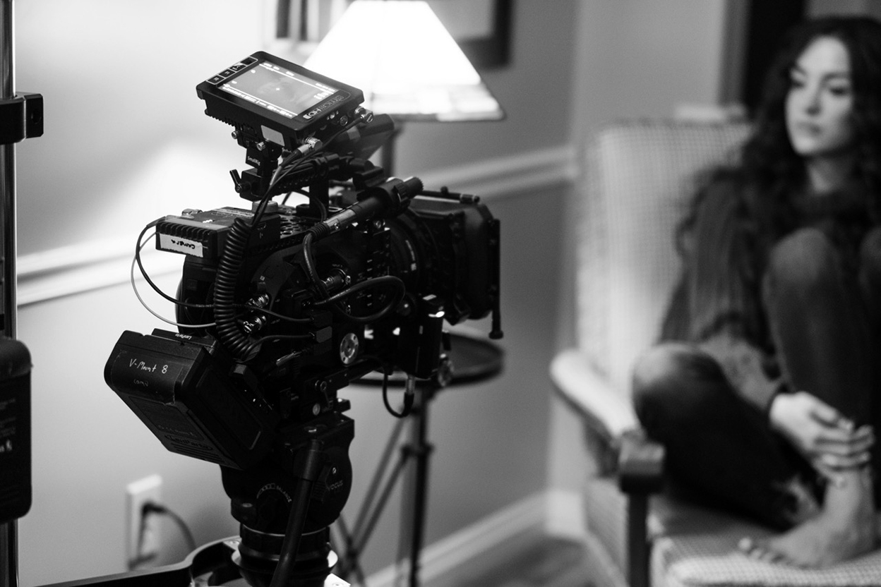 A film camera on set, pointed at an actor sitting in an armchair